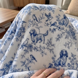 May include: White and blue patterned blanket featuring a toile design with dachshunds, birds, and floral elements. The blanket is soft and plush, with a detailed print. The color scheme is a classic blue and white, creating a cozy and decorative aesthetic.