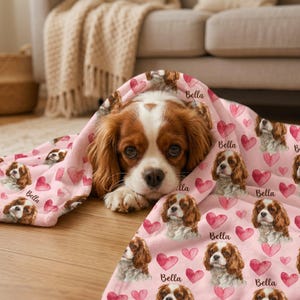 May include: A Cavalier King Charles Spaniel dog peeks out from under a pink blanket. The blanket is decorated with hearts and dog portraits, with the name "Bella" printed on it. The dog has brown and white fur.