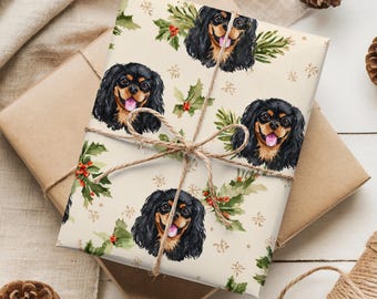 May include: Gift wrapped in cream-colored paper featuring a repeating pattern of Cavalier King Charles Spaniel dog portraits, holly, and snowflakes. The package is tied with twine and rests on a neutral surface with pinecones.