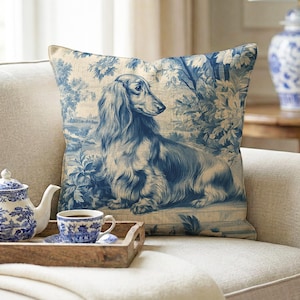 May include: A decorative throw pillow with a blue and white toile design featuring a long-haired dachshund dog. The pillow is on a beige sofa, with a matching teapot, teacup, and serving tray.