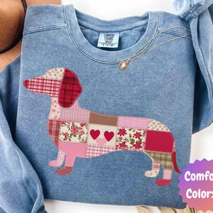 May include: A light blue Comfort Colors sweatshirt featuring a patchwork dachshund design in shades of red, pink, and brown. The dog has two hearts and floral patterns. A gold necklace and a brown hat are also visible.