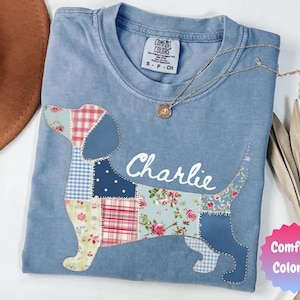 May include: A light blue Comfort Colors t-shirt featuring a patchwork dachshund design with the name "Charlie" in white script. The dog's body is made of various patterned fabrics. A gold necklace with a pendant rests on the shirt.