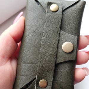 May include: Olive green leather card holder with a flap closure secured by three brass-colored snap buttons. The card holder has a textured surface and a folded design.