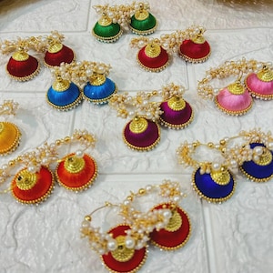 May include: A collection of colorful, handcrafted earrings. The earrings feature a round, silk-wrapped base in various colors, adorned with gold-colored accents and small pearls. The earrings are arranged on a white surface.