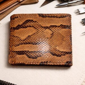 May include: A brown bi-fold wallet with a snakeskin pattern. The wallet is made of leather and has a textured surface. Leatherworking tools are in the background.