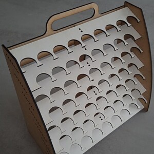 Pode incluir: Um suporte de pintura de madeira com alça, projetado para segurar várias fileiras de garrafas de tinta. O suporte tem um interior branco com recortes circulares para as garrafas.