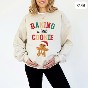 May include: Sand-colored sweatshirt with the text "BAKING a little COOKIE" in red, green, and orange. A gingerbread man wearing a Santa hat is below the text. The sweatshirt is a casual, festive garment.