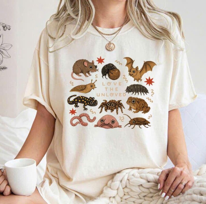 May include: Cream-colored t-shirt featuring a collection of illustrated creatures, including a mouse, bat, and spider. The phrase "LOVE THE UNLOVED" is printed in the center. The shirt has short sleeves and a relaxed fit.