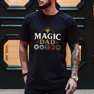 May include: Black t-shirt with the text "MAGIC DAD" in white and gold, with a crown design above the text. Below the text are six colored symbols. The shirt is short-sleeved.