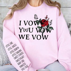 May include: A light pink sweatshirt with the text "I VOW YOU VOW WE VOW" and a red rose graphic with butterflies. The left sleeve has a list of terms including "LITTLE ONE" and "DOLL FACE".