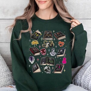 May include: Dark green sweatshirt featuring a collection of illustrated books, skulls, and other fantasy-themed graphics. The design includes titles like "God Fury" and "Hunt the Villain". A unique and stylish apparel item.