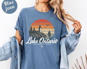 May include: A blue t-shirt featuring a graphic of a lighthouse on a rocky shore, a sailboat, and birds against a sunset circle. The text reads Lake Ontario The Great Lakes with an anchor icon below. The shirt is worn with a blue denim jacket.