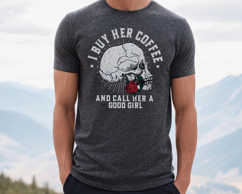 May include: Dark gray t-shirt featuring a skull graphic with a red rose and the text "I BUY HER COFFEE AND CALL HER A GOOD GIRL" in white. The shirt is made of a soft material and has short sleeves.