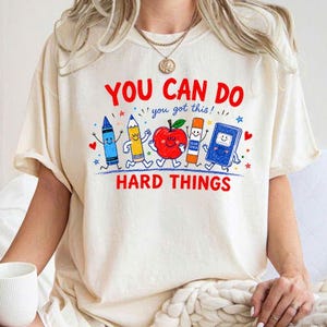 May include: A cream-colored t-shirt featuring the red text "YOU CAN DO HARD THINGS" and the blue text "you got this!". The graphic includes cartoon illustrations of a blue crayon, a yellow pencil, a red apple, a glue stick, and a blue book.