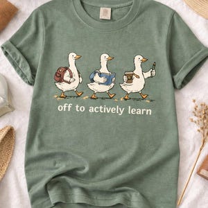 May include: Sage green t-shirt featuring three cartoon ducks walking in a row, each carrying school supplies. The text "off to actively learn" is printed below the ducks. A casual, comfortable tee.