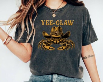 May include: A charcoal gray short-sleeve t-shirt featuring a graphic of a crab wearing a cowboy hat with a star emblem. The text YEE-CLAW is printed in gold above the crab illustration. The shirt has a crew neckline and a vintage-style washed finish.