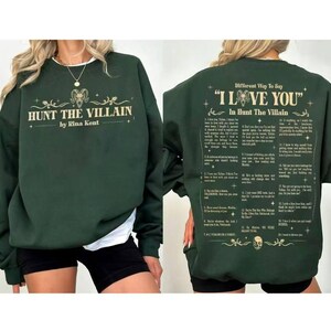 May include: A dark green sweatshirt featuring the text "HUNT THE VILLAIN by Rina Kent" on the front. The back of the sweatshirt displays the text "Different Way To Say 'I LOVE YOU' In Hunt The Villain".