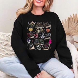 May include: Black sweatshirt featuring a collection of illustrated books, a skull, a flower, a gemstone, a bust, a guitar, and other fantasy-themed elements. The design includes various colors and details, creating a whimsical and artistic aesthetic. The sweatshirt is a casual garment.