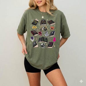 May include: Olive green t-shirt featuring a collection of illustrated books, a skull, a flower, and other graphics. The design includes a variety of colors and shapes, creating a unique and artistic aesthetic. The shirt is a comfortable, casual style.