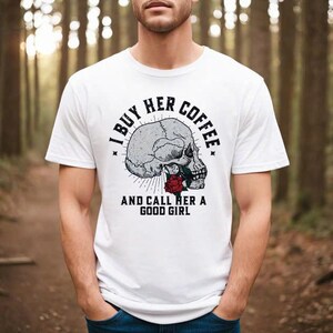 May include: White t-shirt featuring a skull graphic with a red rose and the text "I BUY HER COFFEE AND CALL HER A GOOD GIRL" in black. The shirt is a crew neck and has short sleeves.
