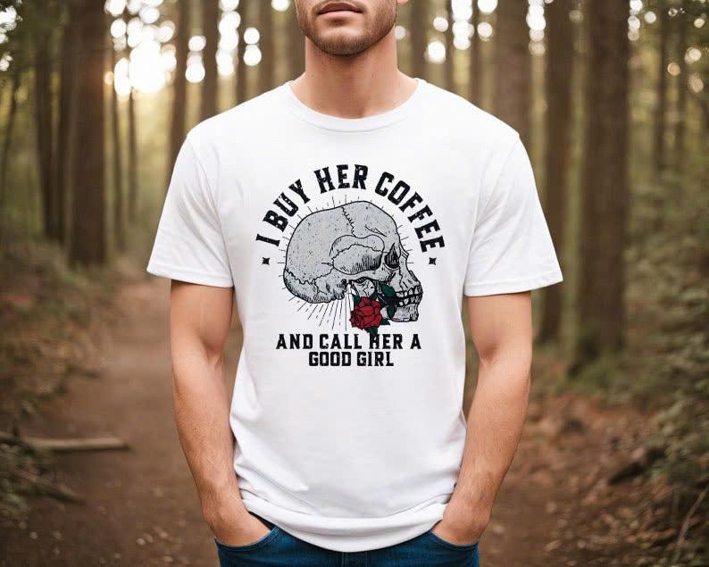 May include: White t-shirt featuring a skull graphic with a red rose and the text "I BUY HER COFFEE AND CALL HER A GOOD GIRL" in black. The shirt is a crew neck and has short sleeves.