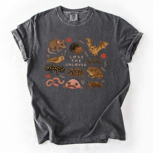 May include: Dark gray t-shirt featuring a colorful illustration of various creatures, including a mouse, bat, and frog. The text "LOVE THE UNLOVED" is printed in the center of the design. The shirt is made of a soft material.