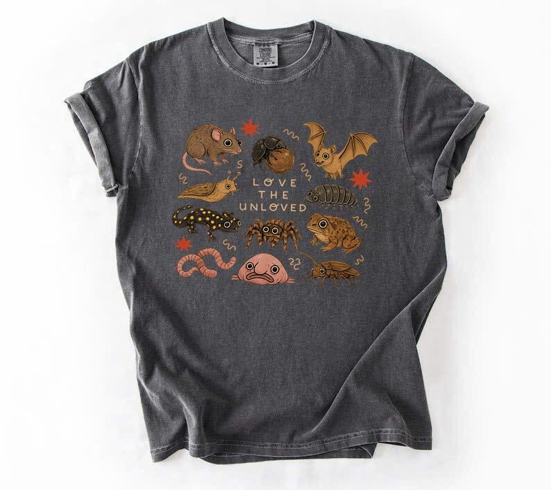 May include: Dark gray t-shirt featuring a colorful illustration of various creatures, including a mouse, bat, and frog. The text "LOVE THE UNLOVED" is printed in the center of the design. The shirt is made of a soft material.