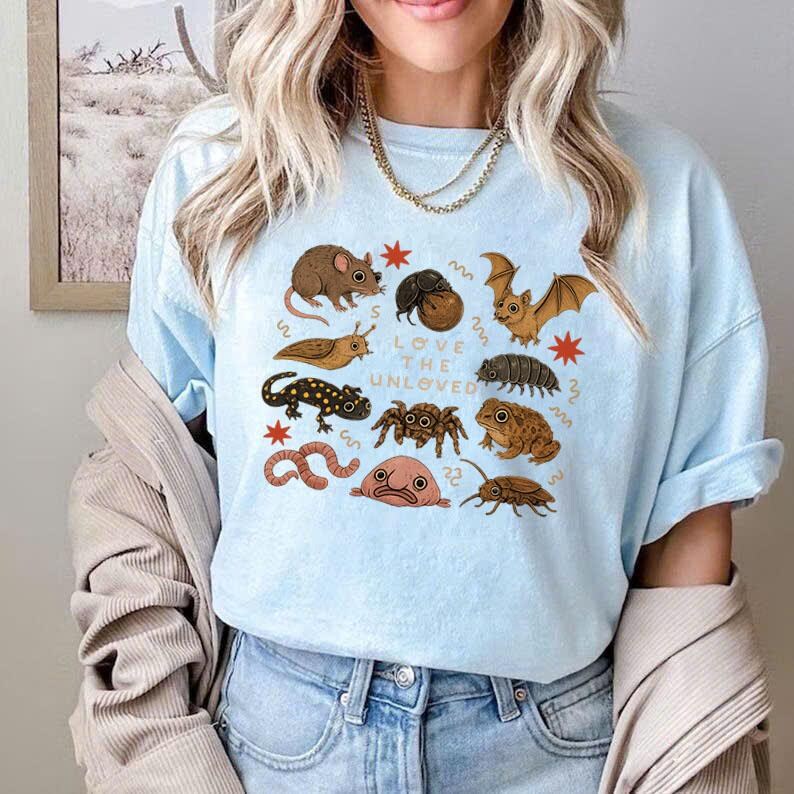 May include: Light blue t-shirt featuring a collection of illustrated insects and small animals, including a bat, spider, and worm. The text "Love the Unloved" is centered on the shirt. The design is in a cartoon style.