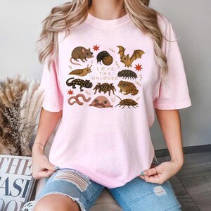 May include: A light pink t-shirt featuring various illustrations of insects and small animals, including a mouse, bat, and spider. The text "LOVE THE UNLOVED" is printed in the center. The shirt is made of a soft material.
