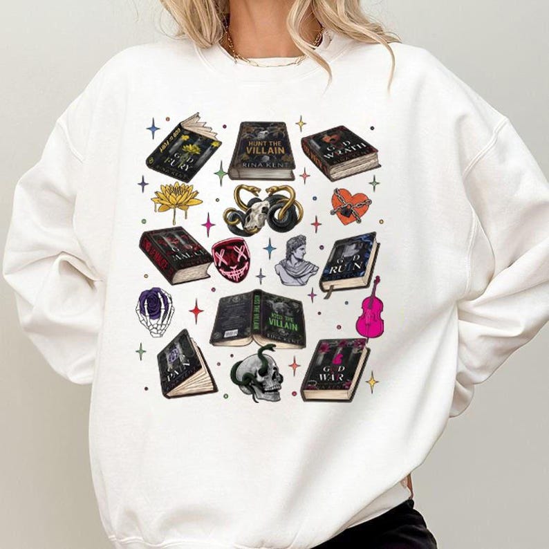 May include: White sweatshirt featuring a collection of illustrated books, a skull, a mask, and other gothic-themed graphics. The books have titles like "Hunt the Villain" and "God Fury". The design includes stars and a pink violin.