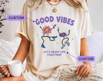May include: A cream-colored t-shirt featuring a purple graphic with the text GOOD VIBES and LET'S ENJOY LIFE TOGETHER. The design includes two dancing flower characters with smiling faces, wearing blue shoes, and holding small bottles.