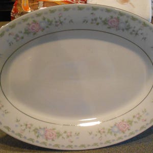 Könnte beinhalten: Ovale Servierplatte mit weißer Oberfläche und floralem Rand. Der Rand zeigt rosa Rosen, blaue Blumen und grüne Blätter. Eine dünne goldene Linie akzentuiert den Rand der Platte.