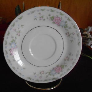 Könnte beinhalten: Weiße Porzellanuntertasse mit einem Blumenmuster mit rosa und blauen Rosen und kleinen grünen Blättern. Die Untertasse hat einen silbernen Rand und wird auf einem goldfarbenen Ständer präsentiert. Ein klassisches Stück für Tee oder Kaffee.
