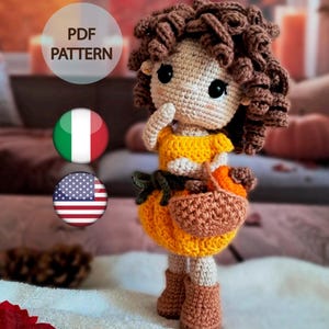 Può includere: Bambola all'uncinetto con capelli ricci castani, indossa un top giallo e una gonna a forma di zucca. La bambola tiene un piccolo cestino e indossa stivali marroni. L'immagine include il testo "PDF PATTERN" e le bandiere italiana e americana.