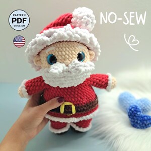 Può includere: Una bambola di Babbo Natale all'uncinetto realizzata con filato rosso, bianco e marrone. La bambola presenta un cappello rosso con un pompon bianco, una barba bianca, occhi blu e una cintura marrone con fibbia gialla. Il testo recita PATTERN PDF ENGLISH e NO-SEW.