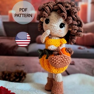 Può includere: Bambola all'uncinetto fatta a mano con capelli ricci castani, che indossa un top giallo e una gonna a forma di zucca. La bambola tiene un piccolo cestino e indossa stivali marroni. L'immagine include il testo "PDF PATTERN" e una grafica della bandiera americana.