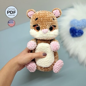 Può includere: Un peluche lavorato all'uncinetto color marrone e crema con accenti rosa su orecchie, mani e piedi. Il giocattolo presenta grandi occhi arancioni, un piccolo naso rosa e sopracciglia ricamate. Un'icona circolare mostra il testo PATTERN PDF ENGLISH.