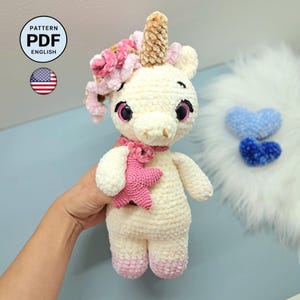 Può includere: Un peluche a forma di unicorno all'uncinetto color crema con un corno dorato testurizzato, un accessorio a forma di stella rosa e una criniera di filato riccio rosa e bianco. Il giocattolo ha grandi occhi scuri e zoccoli rosa. Include l'icona PATTERN PDF ENGLISH.