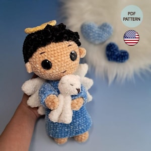 Può includere: Una bambola angelo all'uncinetto fatta a mano con capelli neri, un'aureola gialla e ali bianche. L'angelo indossa un vestito blu e tiene in mano un orsacchiotto bianco. Due decorazioni all'uncinetto a forma di cuore blu sono sullo sfondo.