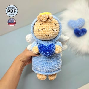 Può includere: Una bambola angelo all'uncinetto con tunica azzurra con cappuccio, ali bianche e aureola gialla. La bambola ha un viso beige con occhi chiusi e tiene in mano un cuore blu scuro testurizzato. Un'icona mostra il testo PATTERN PDF ENGLISH.