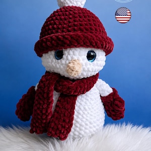 Può includere: Un pupazzo di neve all'uncinetto bianco con un berretto rosso scuro con pompon bianco, una sciarpa rosso scuro abbinata e muffole rosso scuro. Il pupazzo di neve ha grandi occhi blu e un piccolo naso beige. Il testo nell'immagine dice PATTERN PDF ENGLISH.