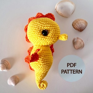 Può includere: Un cavalluccio marino all'uncinetto fatto a mano, giallo e arancione. Il cavalluccio marino ha una testa arrotondata, una coda arricciata e occhi neri. L'immagine include conchiglie e un cerchio blu con il testo "PDF PATTERN".