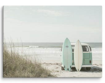 May include: A vintage, light blue Volkswagen van with two surfboards strapped to the back. The van is parked on a sandy beach with ocean waves in the background.