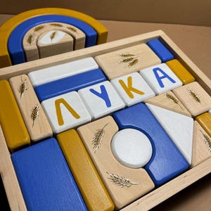 May include: A wooden toy block set with a rainbow arch and blocks painted in blue, yellow, and white. The blocks have the name "LYUKA" spelled out on them and a wheat design.