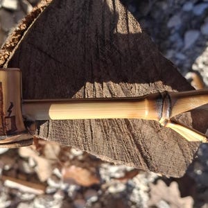 Può includere: Una pipa in bambù fatta a mano con una testa cilindrica con un disegno intagliato. Lo stelo della pipa si estende dalla testa, mostrando la venatura naturale del bambù. La pipa è appoggiata su una superficie di legno stagionato.