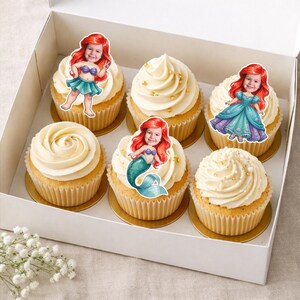 Op de afbeelding: Een witte doos bevat zes cupcakes met vanille glazuur en gouden sprinkles. Elke cupcake is versierd met een cartoon zeemeermin afbeelding met rood haar. De cupcakes staan op gouden cupcake vormpjes.