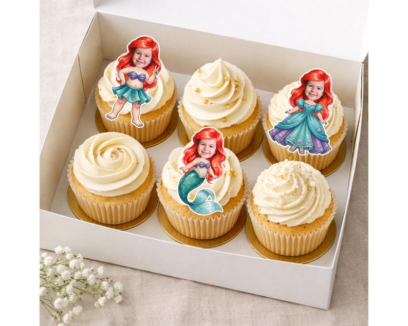 Op de afbeelding: Een witte doos bevat zes cupcakes met vanille glazuur en gouden sprinkles. Elke cupcake is versierd met een cartoon zeemeermin afbeelding met rood haar. De cupcakes staan op gouden cupcake vormpjes.