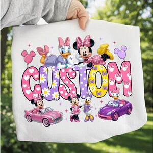 May include: A white t-shirt featuring a colorful graphic of Minnie Mouse and Daisy Duck. The word CUSTOM is printed in large, patterned letters. The design includes illustrations of the characters, a pink convertible, a purple car, and balloons.