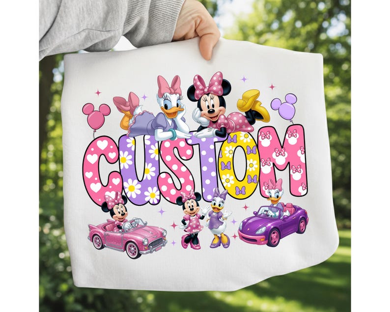 May include: A white t-shirt featuring a colorful graphic of Minnie Mouse and Daisy Duck. The word CUSTOM is printed in large, patterned letters. The design includes illustrations of the characters, a pink convertible, a purple car, and balloons.