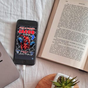 Peut inclure: Un smartphone noir affichant une invitation d'anniversaire Spiderman. L'invitation est pour le 7&egrave;me anniversaire de Jacob et pr&eacute;sente une illustration de Spiderman rouge et bleu. Le t&eacute;l&eacute;phone est connect&eacute; &agrave; un c&acirc;ble de charge blanc.